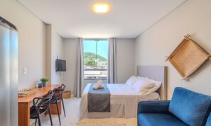 A hotel room with a bed, a desk, a chair, a blue sofa, a TV, and a window with a view of a hill. - Complete Studio 650m From the Beach - BC - Dpr0003 (Balneário Camboriú)