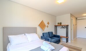 A hotel room with a bed, a blue chair, a small table, and a wall-mounted rack.
