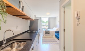 A compact hotel room with a kitchenette, a bed with a blue headboard, a bedside table, a telephone, and a view of the outside through a window. - Complete Studio 650m From the Beach - BC - Dpr0003 (Balneário Camboriú)