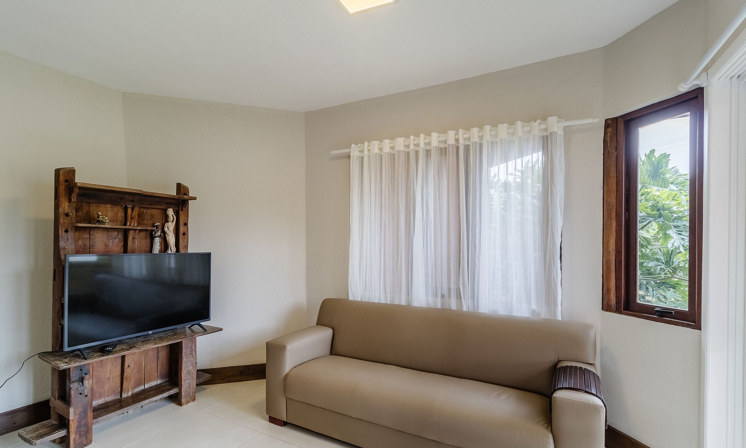 A living room with a flat-screen TV on a wooden stand, a beige sofa, and a window with sheer curtains.