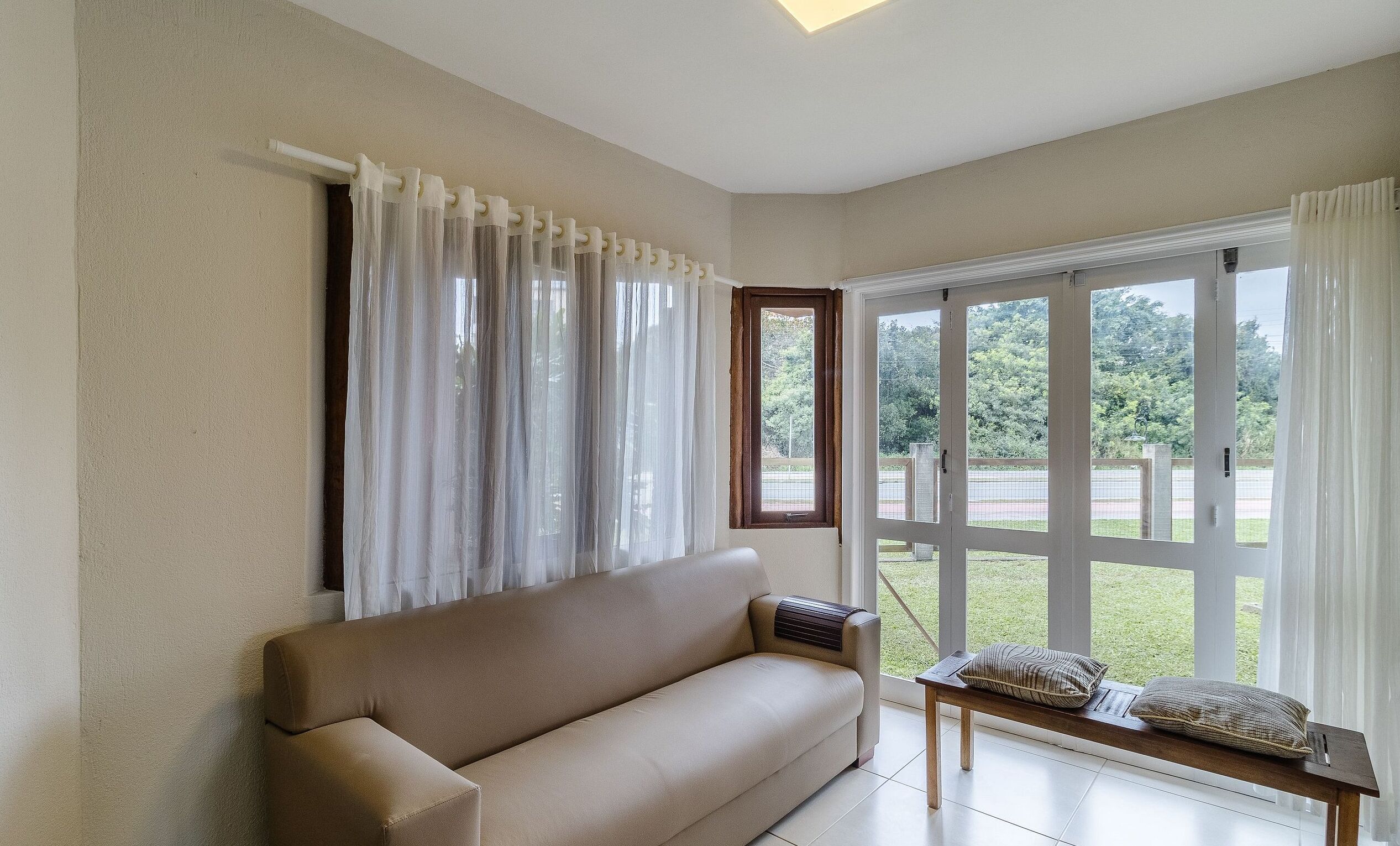 A room with a beige sofa, a small table with cushions, a window with curtains, and a view of greenery outside.