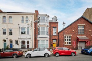 Une vue de rue avec des bâtiments anciens, dont une maison d’angle dotée d’une baie vitrée et un bâtiment en briques rouges sur lequel on peut lire l’inscription « Saltburn Vintage Rooms ».