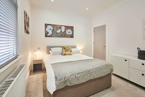 Une chambre à coucher avec un lit, des lampes de chevet, un radiateur, une porte et un mur orné d’un tableau représentant des motifs floraux.