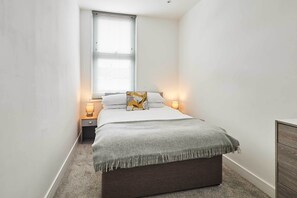 Ein schmaler Raum mit einem Bett, zwei Nachttischlampen, einem Fenster mit Jalousien und einer Heizung.