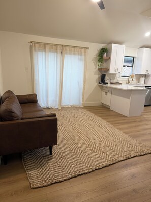 A modern living room with a brown sofa, a beige rug, and a kitchen area with white cabinets and a sink. - Cozy Modern 3-bedroom house with AC in tranquil Fort Worth (Fort Worth)