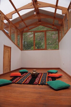 A room with a sloped ceiling, large windows, a wooden door, and a patterned rug with cushions. - A beautiful private home surrounded by mountains and trails (Urubamba)