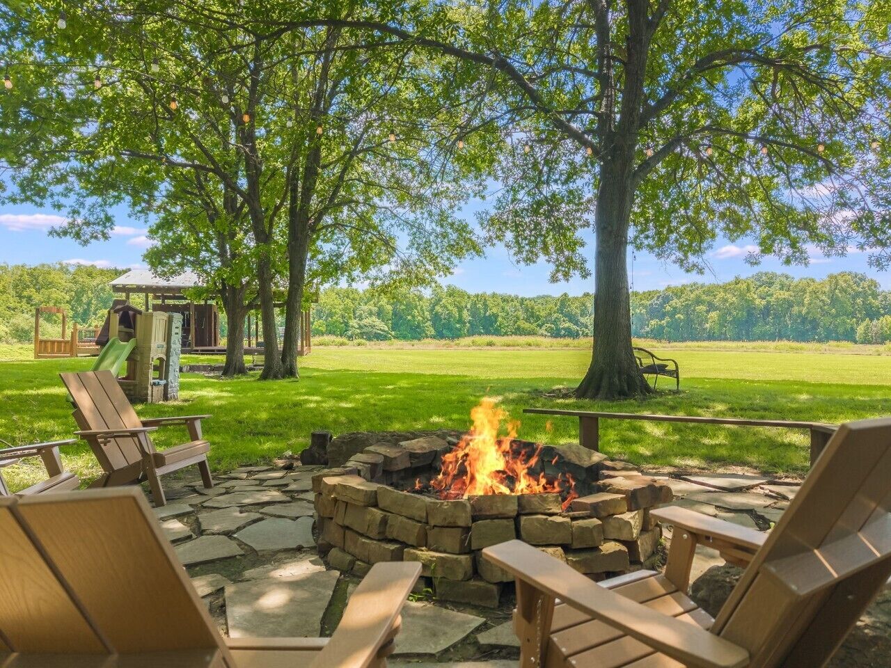 A fire pit with seating, surrounded by trees and a playground in a grassy area.