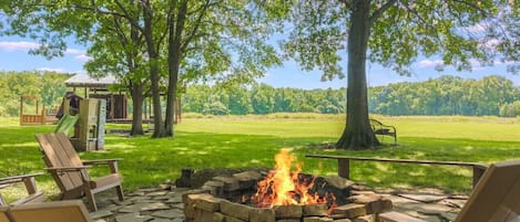 A fire pit with seating, surrounded by trees and a playground in a grassy area.