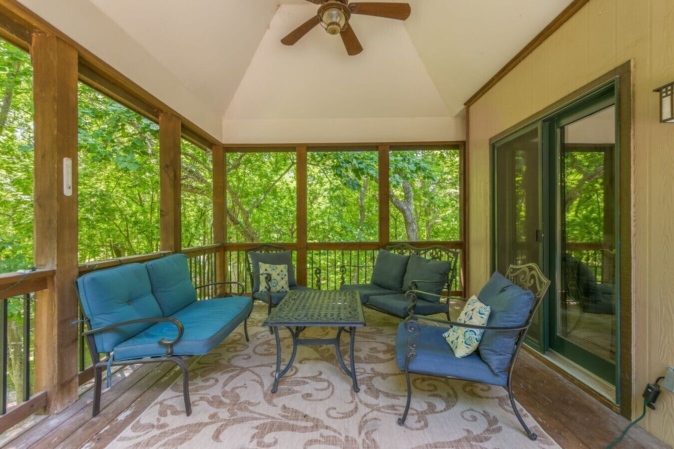 A covered porch with a ceiling fan, wooden beams, and a view of green trees.