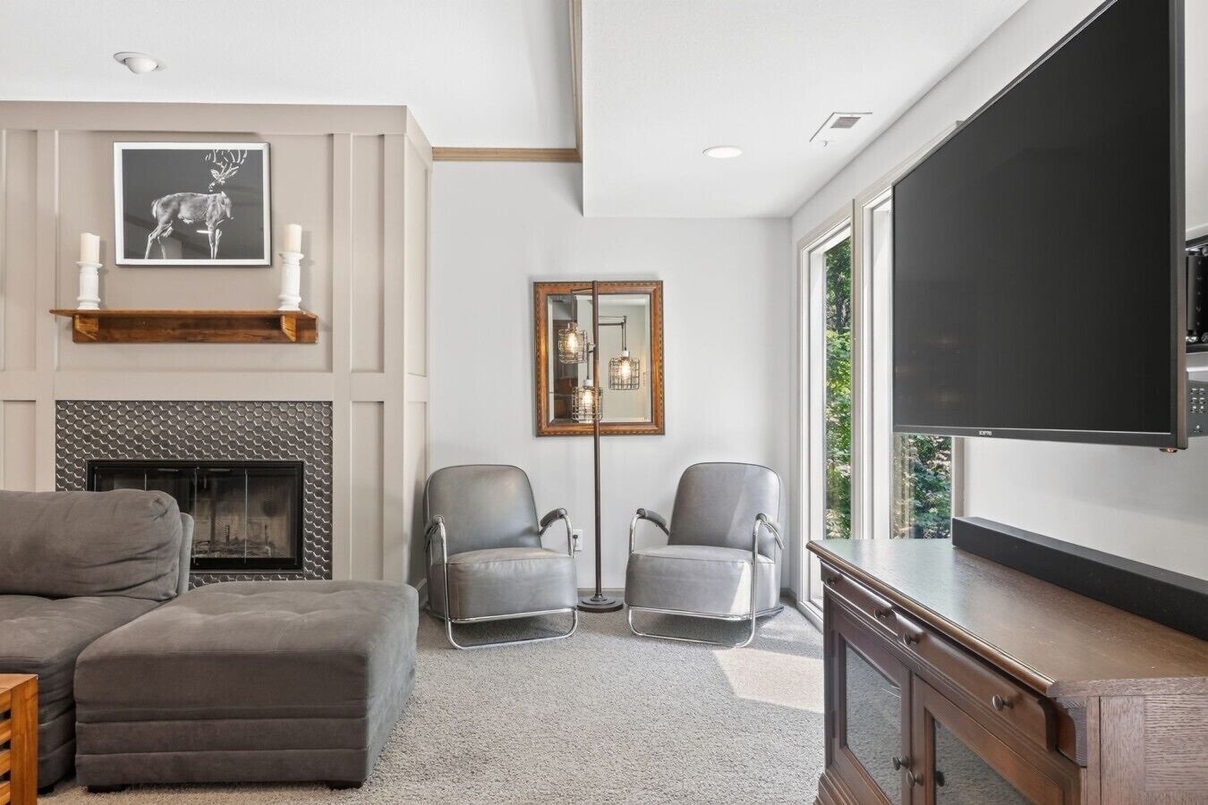 A modern living room with a fireplace, two grey armchairs, a sofa, and a large flat-screen TV mounted on the wall.