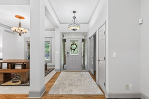A hallway with a wooden floor, a rug, a door with a wreath, and a chandelier.
