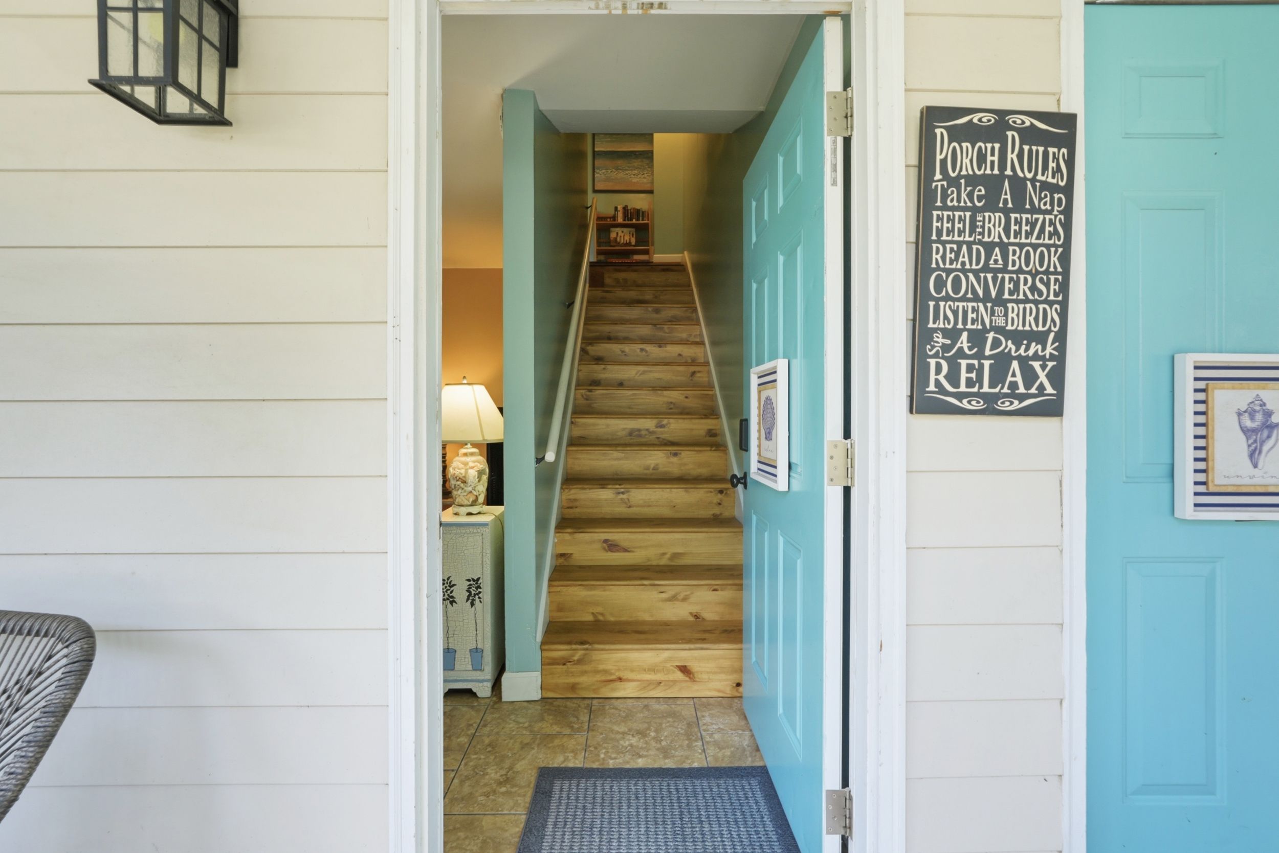 A wooden staircase leading to a room with a teal door, a sign with porch rules, and a lamp on a nightstand.