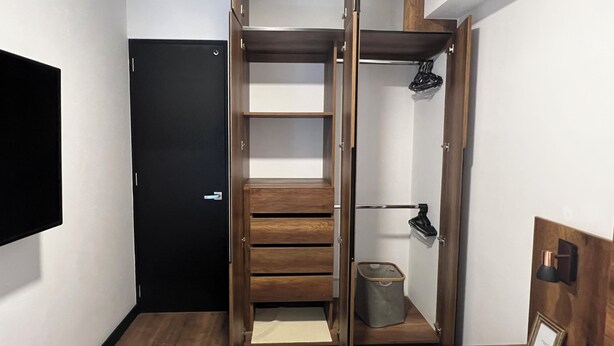 Quarto compacto com roupeiro embutido, uma pequena lixeira e luminária fixada na parede.