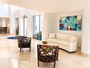 Un hall d’entrée spacieux, avec un canapé blanc, deux fauteuils en bois, une table basse et un tableau abstrait aux couleurs vives accroché au mur.