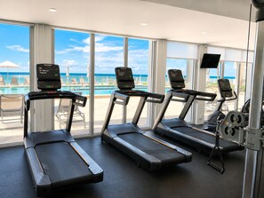 Une salle de sport équipée de tapis de course, d’un banc de musculation et offrant une vue sur une piscine et une plage.