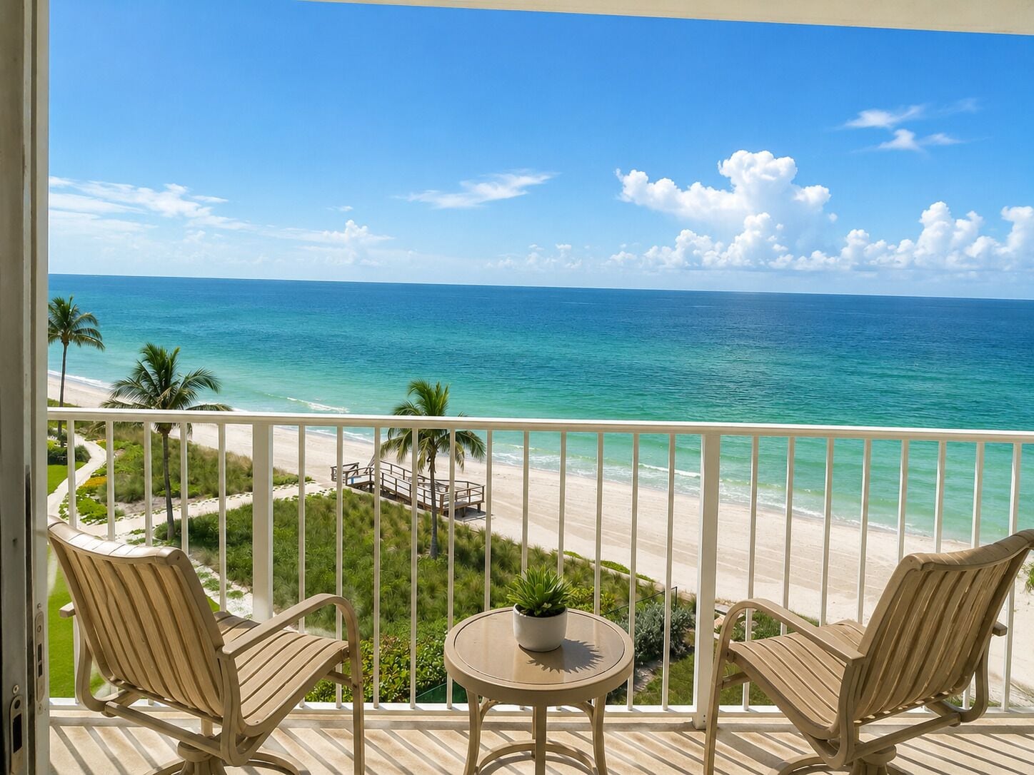 A balcony with two chairs and a small table, overlooking a beach with palm trees and a clear blue sky.