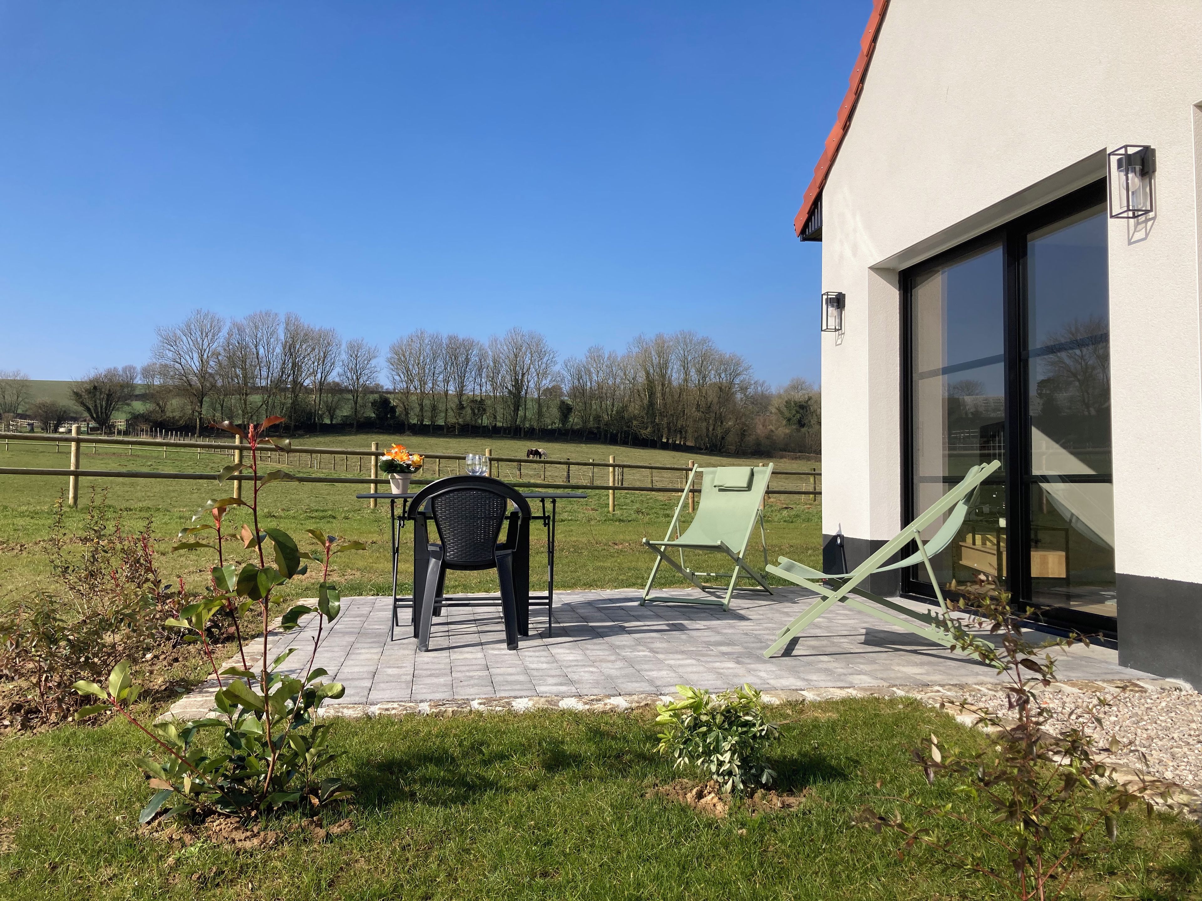 Une terrasse avec une table et des chaises, un petit jardin et une vue sur un champ clôturé planté d’arbres.