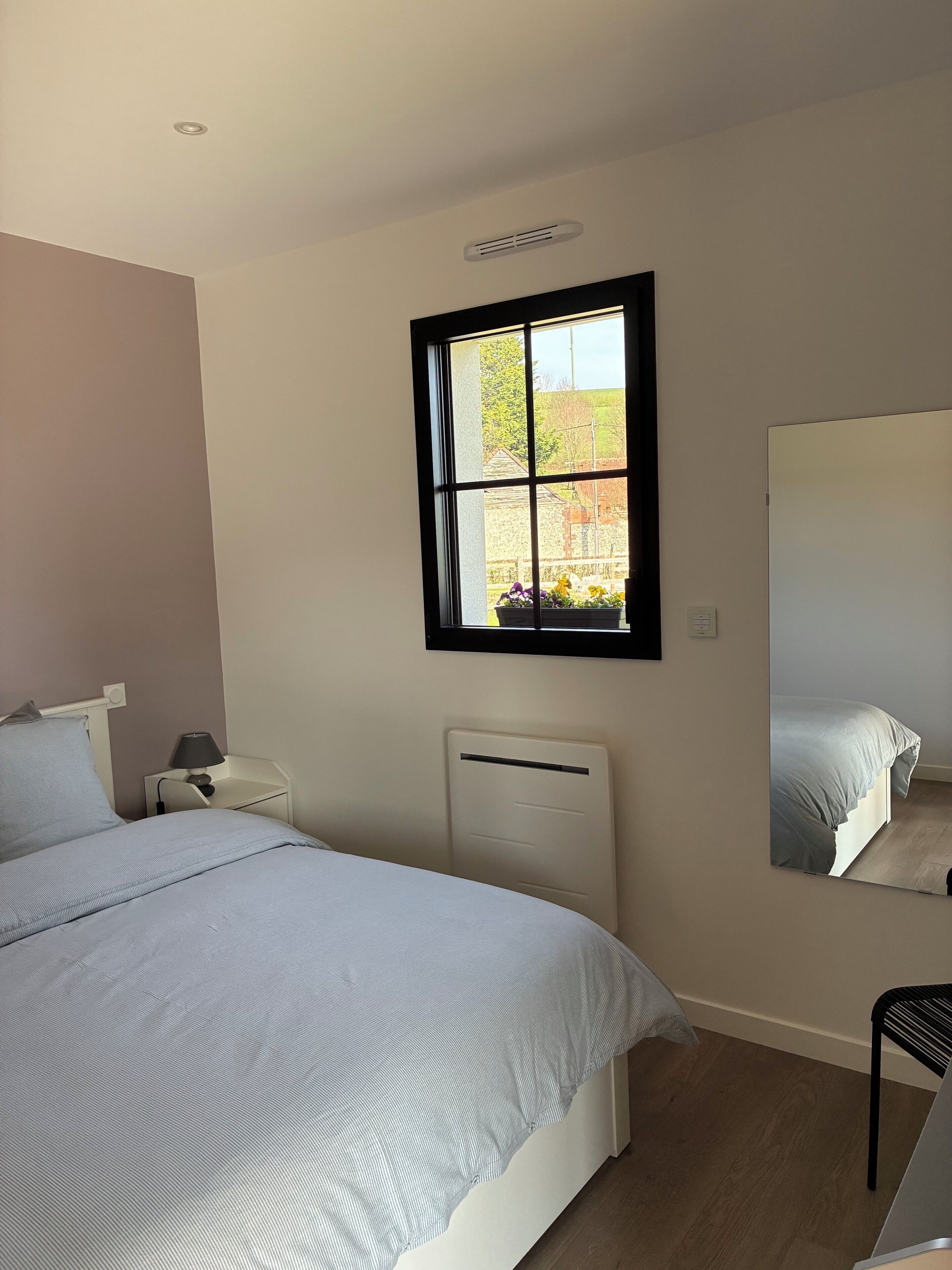 Ein Schlafzimmer mit einem Bett, einem Nachttisch, einer Lampe, einer Heizung, einem Fenster und einem Spiegel.