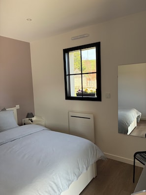 Ein Schlafzimmer mit einem Bett, einem Nachttisch, einer Lampe, einer Heizung, einem Fenster und einem Spiegel.