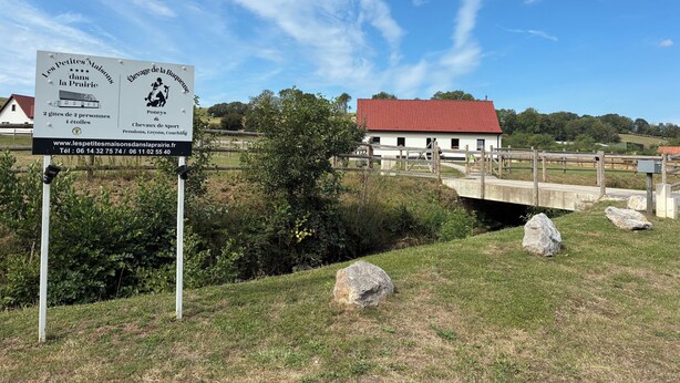 Ein Schild mit Informationen zu Unterkünften und Dienstleistungen, ein weißes Gebäude mit rotem Dach und eine Holzbrücke in ländlicher Umgebung.