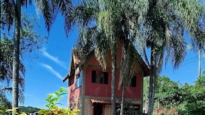 A house with a red exterior, a porch with wooden chairs, and a small garden area with gravel and greenery. - Cabanas VillaRejo (Jarinu)