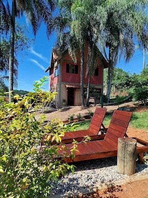 Una casa con fachada roja, un porche con sillas de madera y un pequeño jardín con grava y vegetación.