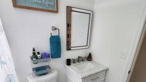 A bathroom with a toilet, sink, mirror, and a towel rack. - Be A Nomad | Cozy 2br Home Steps from the Beach (Jacksonville Beach)
