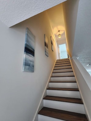 Eine Treppe mit Holzhufen, die nach oben führt, begrenzt von weißen Wänden und gerahmten Kunstwerken.