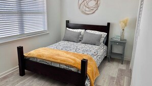 A bedroom with a bed, a bedside table, a wall decoration, and a window with blinds. - Be A Nomad | Cozy 2br Home Steps from the Beach (Jacksonville Beach)