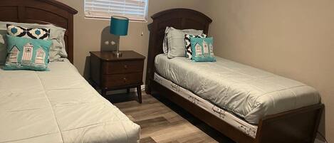 A bedroom with two single beds, a nightstand, a lamp, and a window with blinds.
