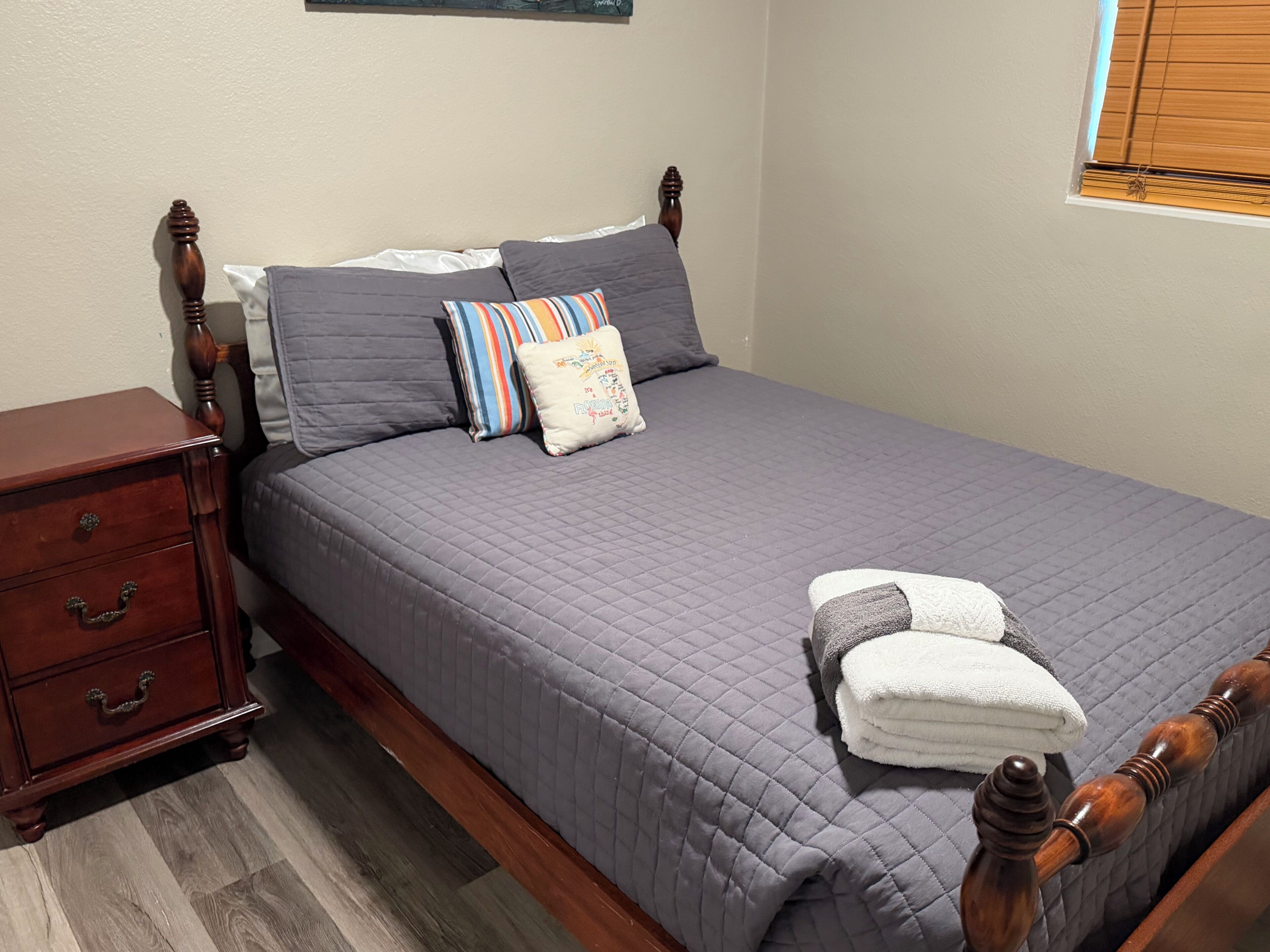 A neatly made bed with a grey quilt, a wooden nightstand with drawers, and a window with blinds.
