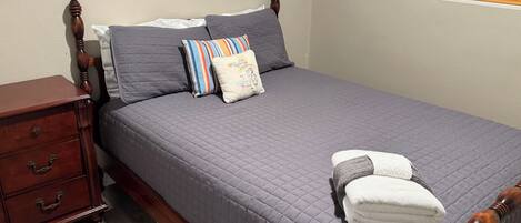 A neatly made bed with a grey quilt, a wooden nightstand with drawers, and a window with blinds.