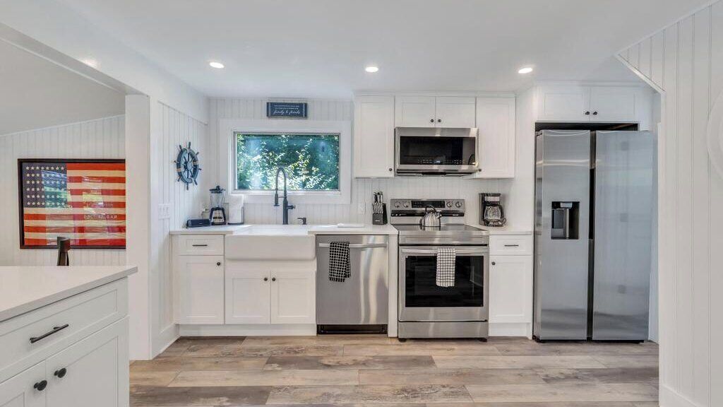 A modern kitchen with white cabinetry, stainless steel appliances, a large window with a view, and a mounted clock.