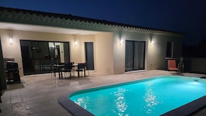 Una piscina con bordo piastrellato, zona pranzo con sedie e patio coperto con una poltrona.