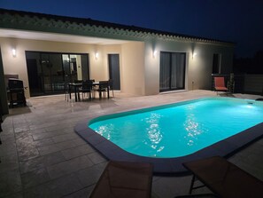 Une piscine avec une terrasse carrelée, un coin repas avec des chaises et un patio couvert avec un fauteuil.