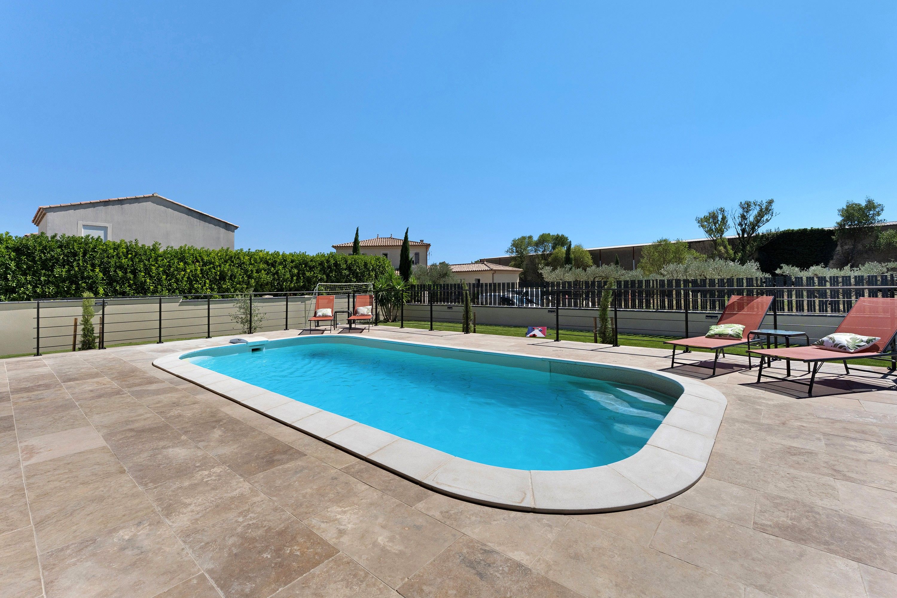 Une piscine rectangulaire avec une terrasse carrelée, entourée d’une petite clôture et d’une haie.