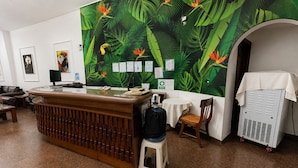 A reception area with a wooden counter, a white stool, a small table, and a wall with a tropical leaf pattern. - Gran Hotel Mediterráneo (Santa Cruz de la Sierra)