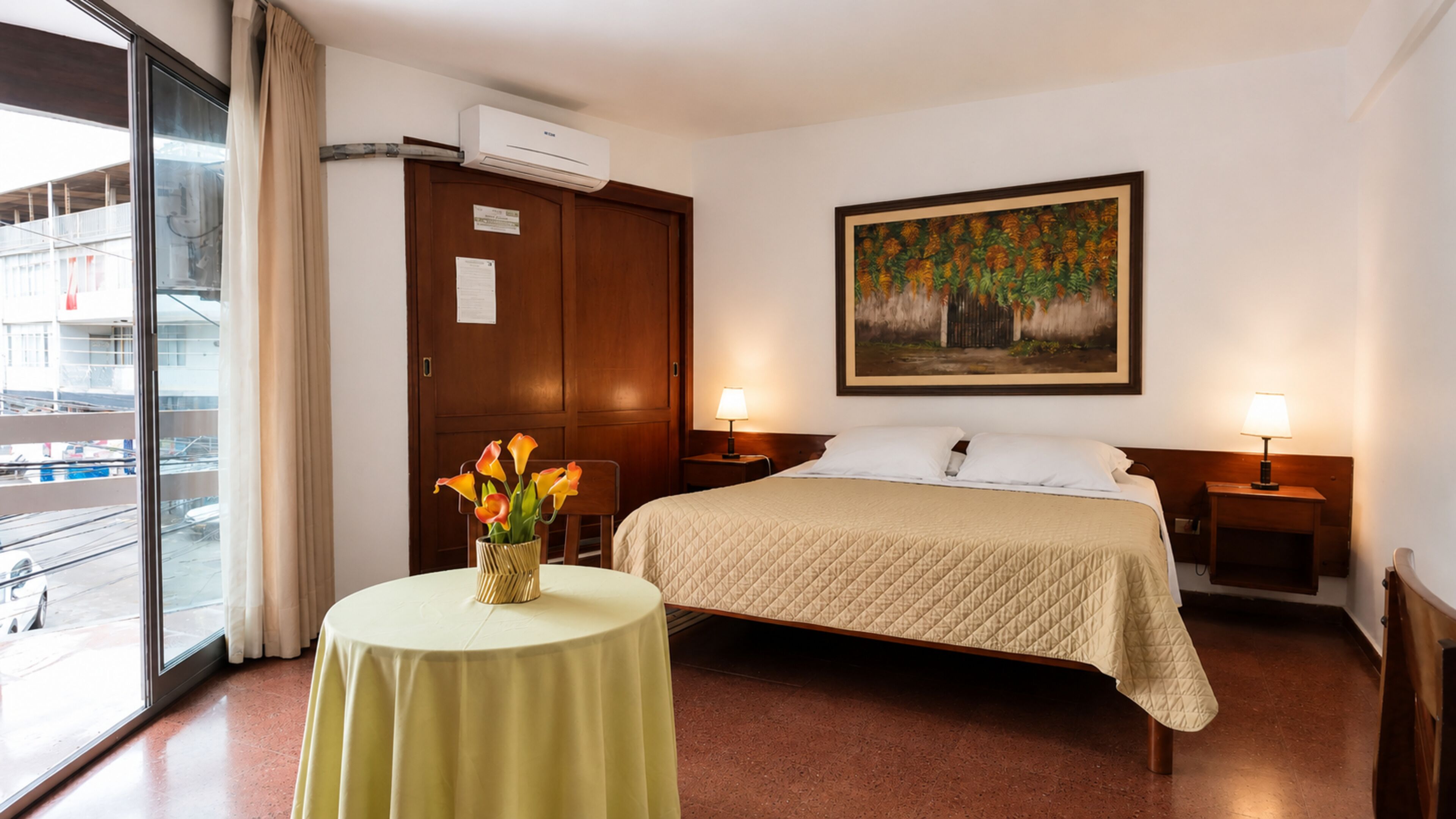 Quarto de hotel com cama, duas luminárias de cabeceira, um quadro na parede, uma janela com vista para o exterior e uma pequena mesa com um vaso de flores.