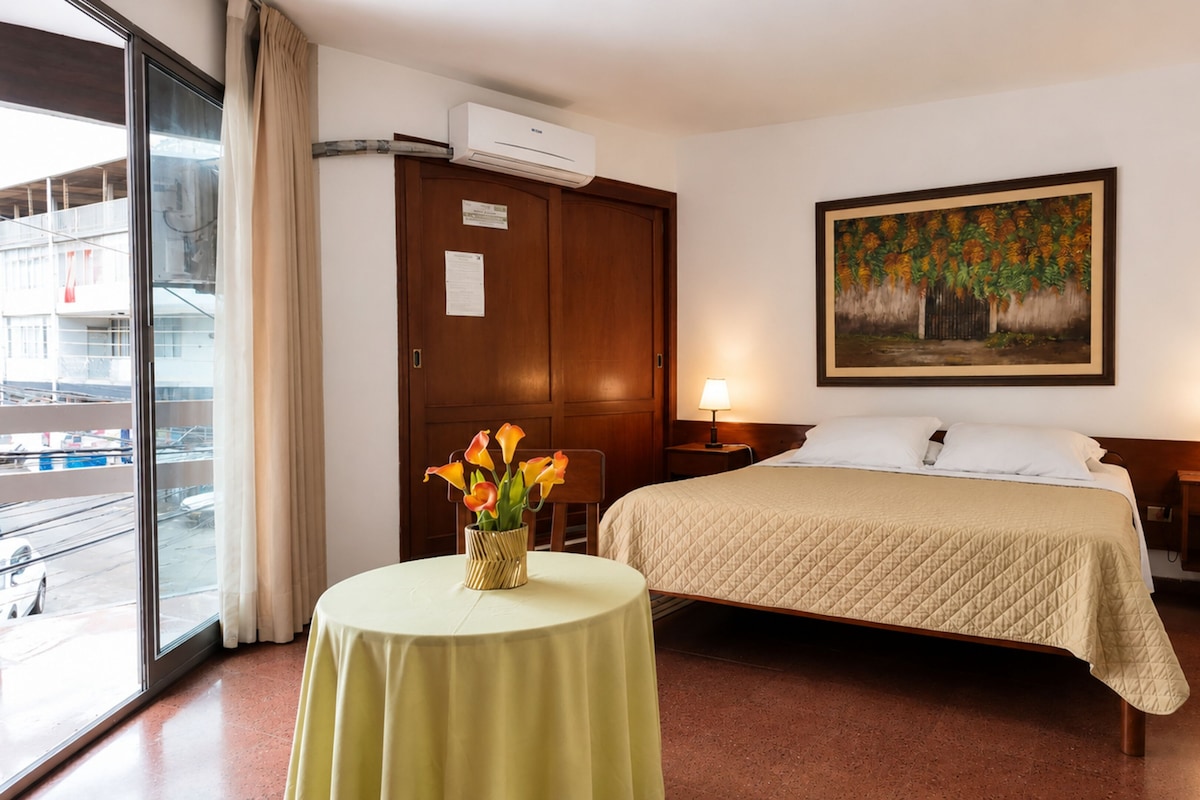 Quarto de hotel com cama, duas luminárias de cabeceira, um quadro na parede, uma janela com vista para o exterior e uma pequena mesa com um vaso de flores.
