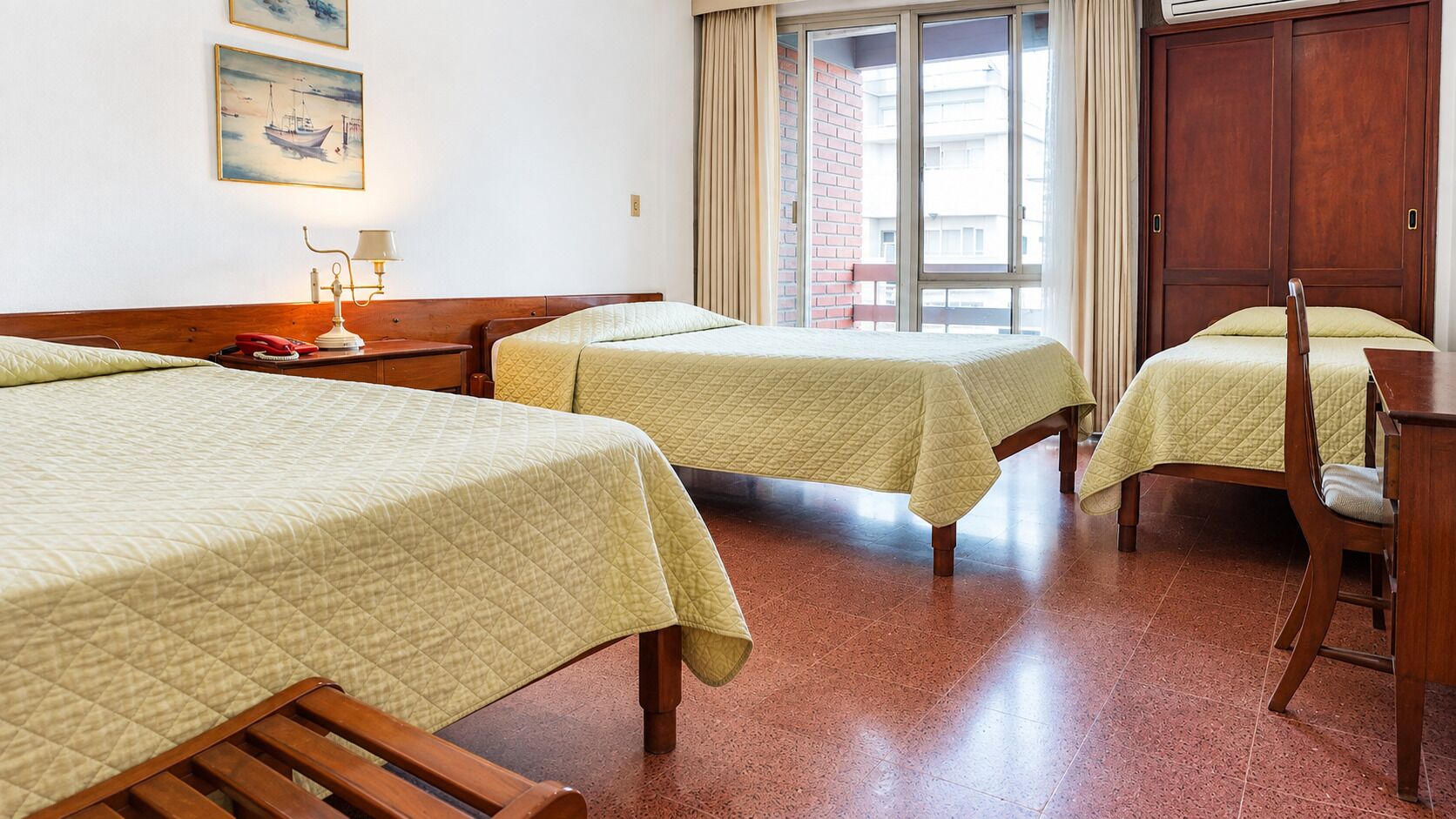 Une chambre d’h ôtel avec trois lits simples, une table de chevet en bois, une lampe et une fenêtre avec des rideaux.