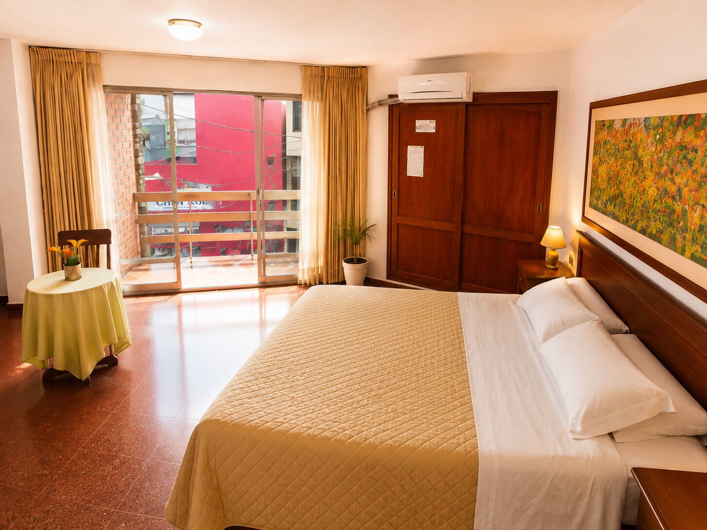 Une chambre d’hôtel avec un lit, une table avec une lampe, une plante en pot et une porte-fenêtre coulissante donnant sur l’extérieur.
