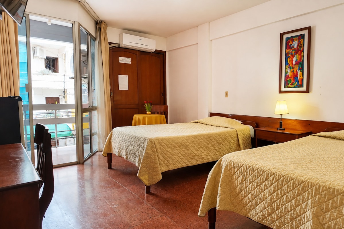 Quarto de hotel com duas camas, uma mesa, uma cadeira, aparelho de ar condicionado e um quadro na parede.