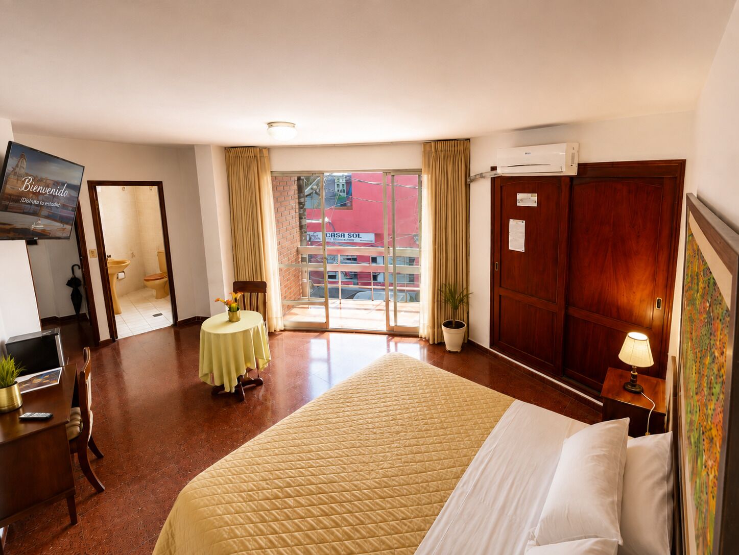 A hotel room with a bed, a desk, a chair, a small table with a vase, a potted plant, and a television on the wall.