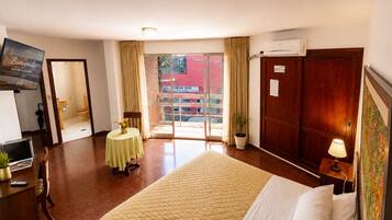 A hotel room with a bed, a desk, a chair, a small table with a vase, a potted plant, and a television on the wall.