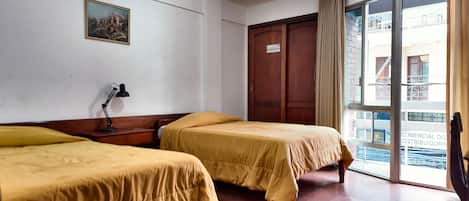 A hotel room with two beds, a desk with a lamp, a large window, and a door.