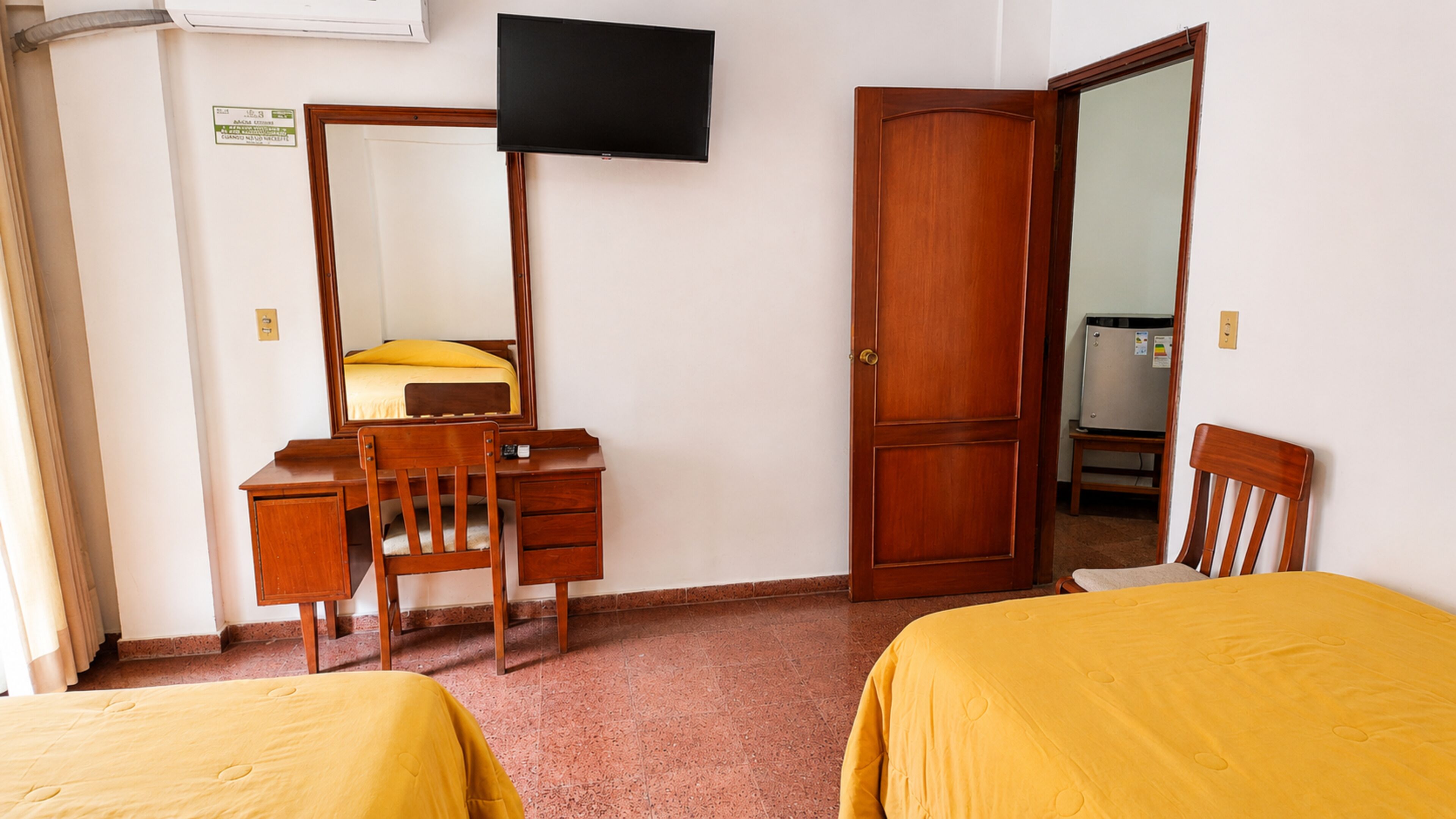 Una camera d'albergo con una scrivania in legno, una sedia, un letto con lenzuola gialle, una televisione a muro e un armadio.