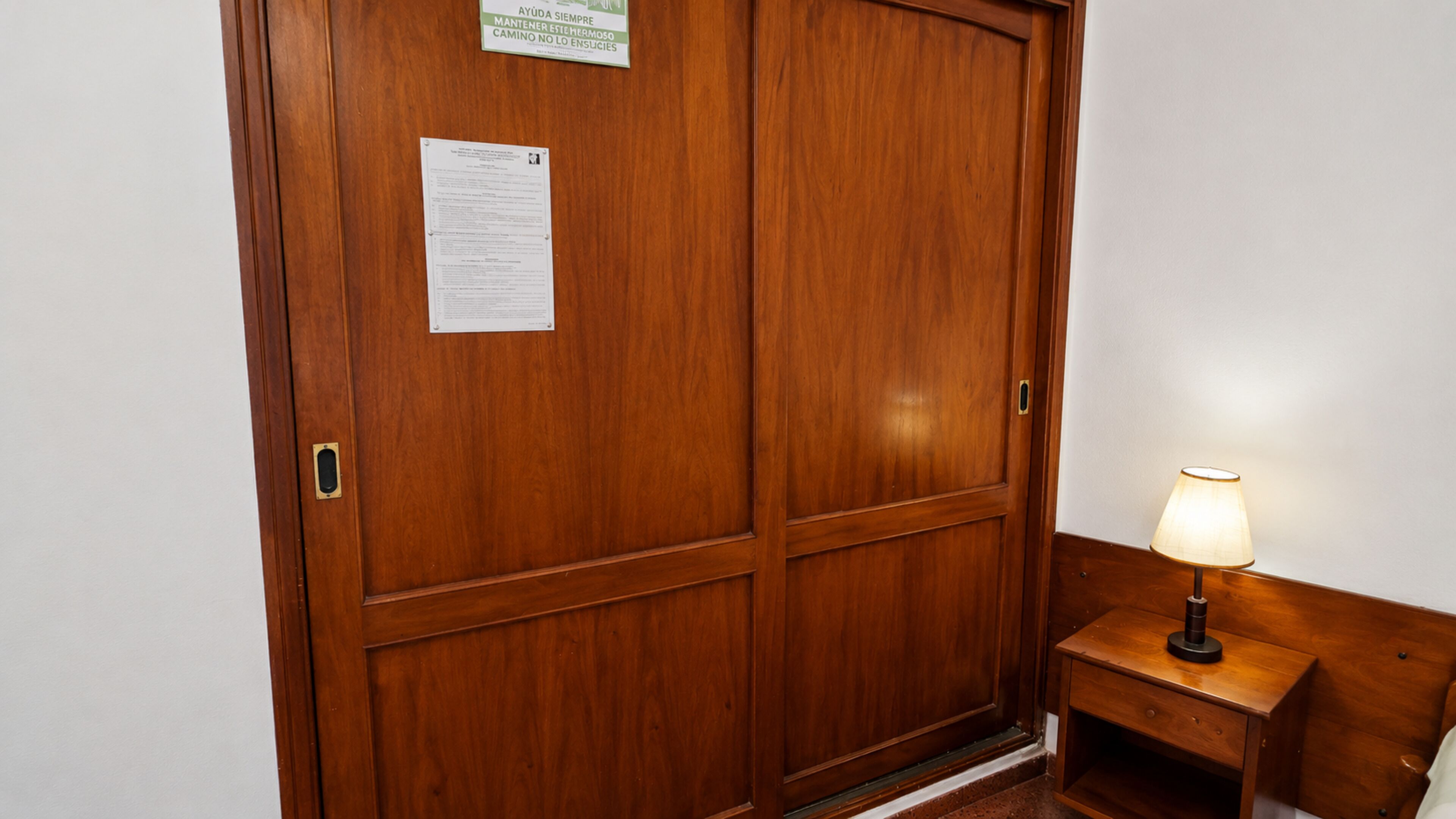 A wooden door with a notice and a small bedside table with a lamp.
