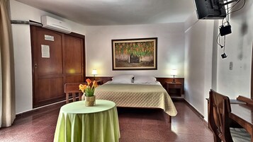 A hotel room with a bed, bedside tables, a desk with a chair, a table with a vase of flowers, and a painting on the wall.