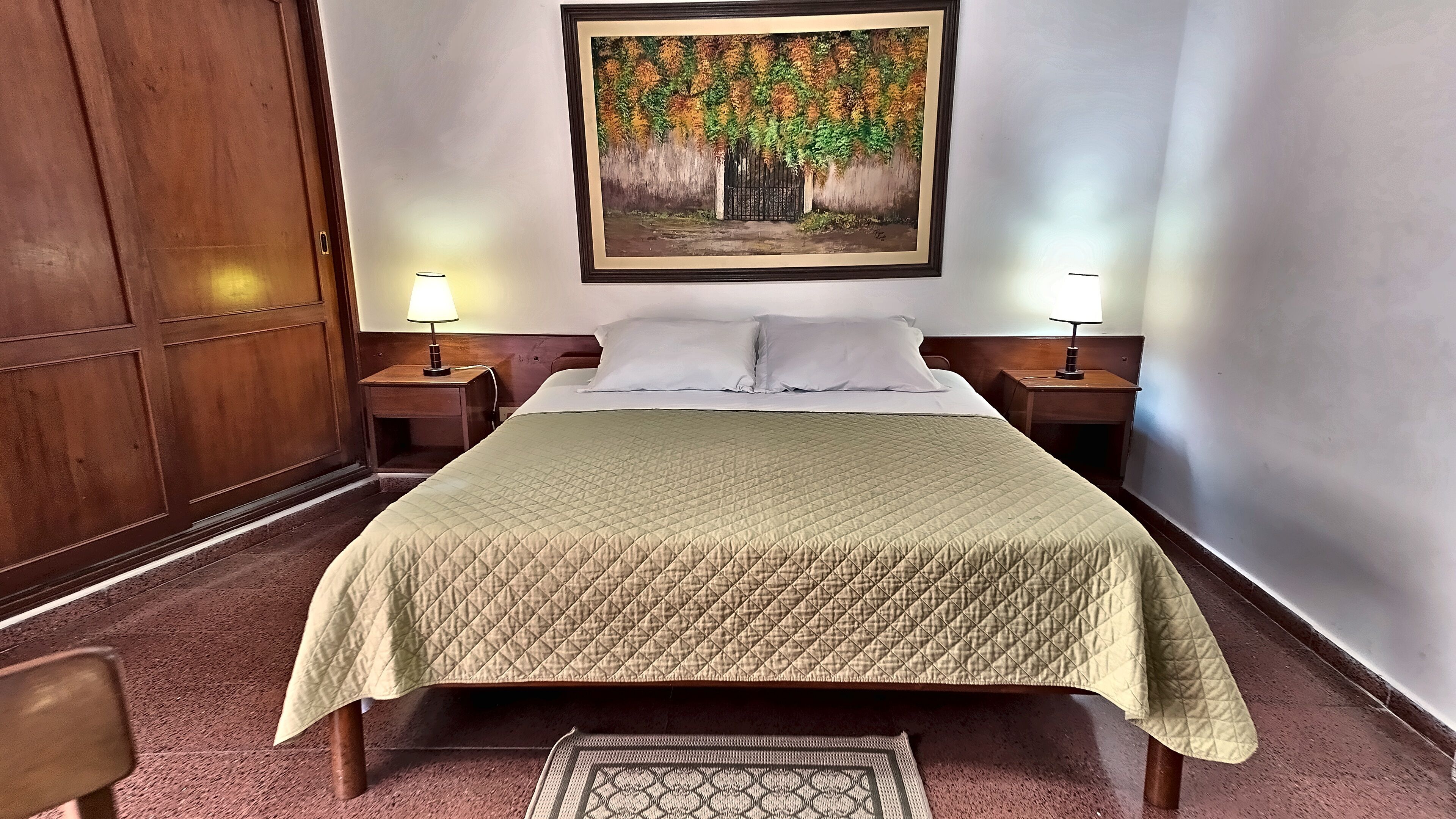 A hotel room with a bed, two bedside tables with lamps, a large painting on the wall, and a wooden wardrobe.