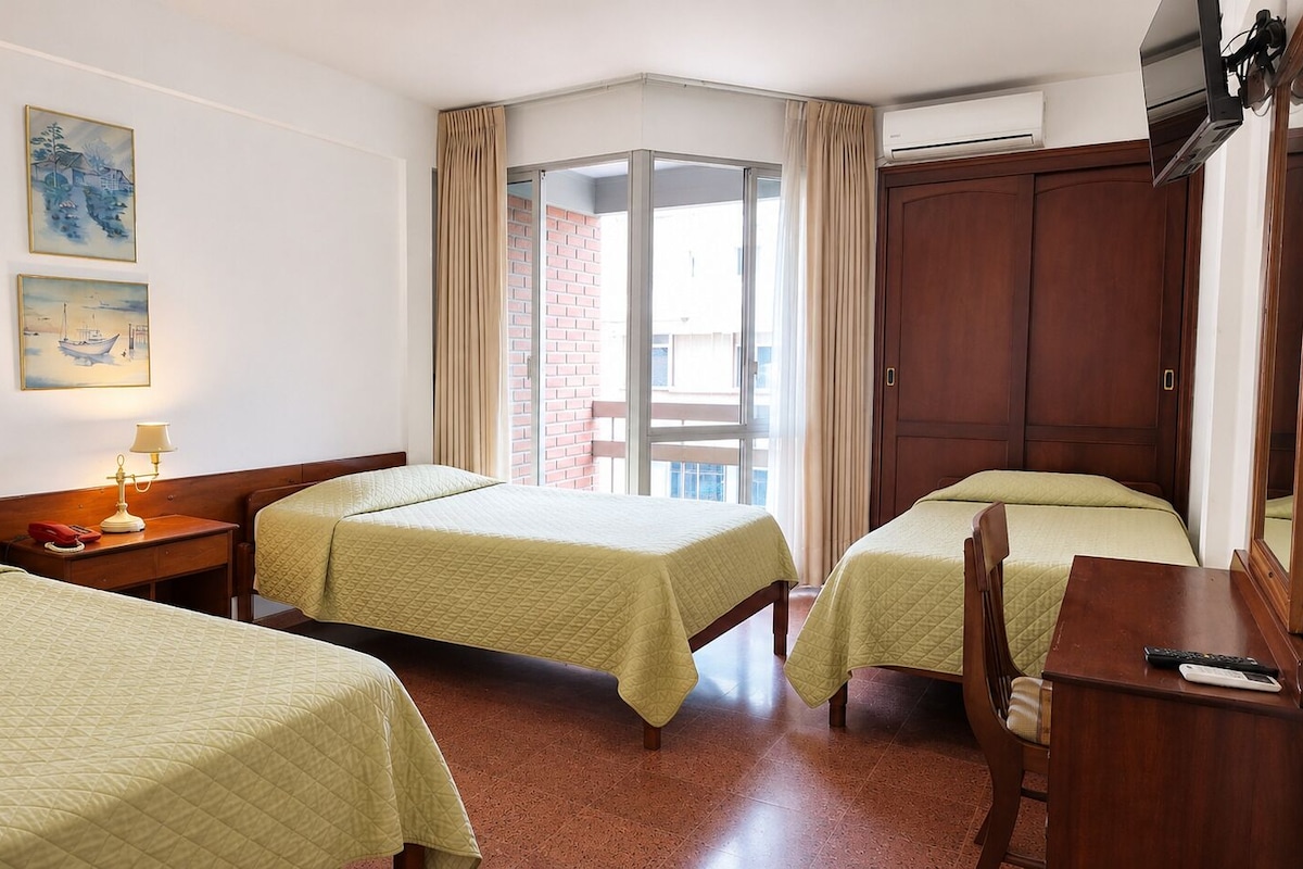 Quarto de hotel com duas camas, uma escrivaninha, uma cadeira, um abajur e uma porta de vidro deslizante que dá para uma área externa.